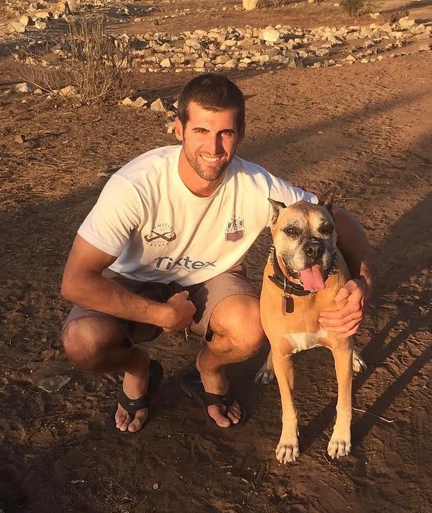 Man kneeling next to a dog in a dry outdoor area.
