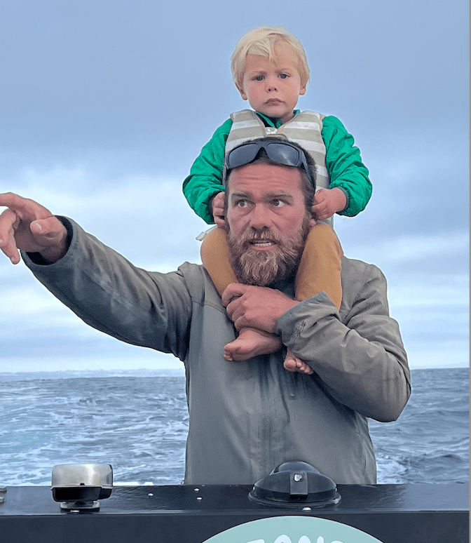 Man carrying child on shoulders, pointing out at sea on a cloudy day.