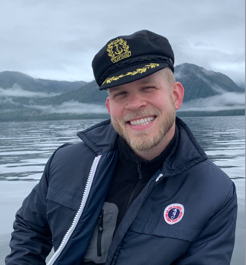 Smiling man in captain's hat on a boat with misty mountains in the background.