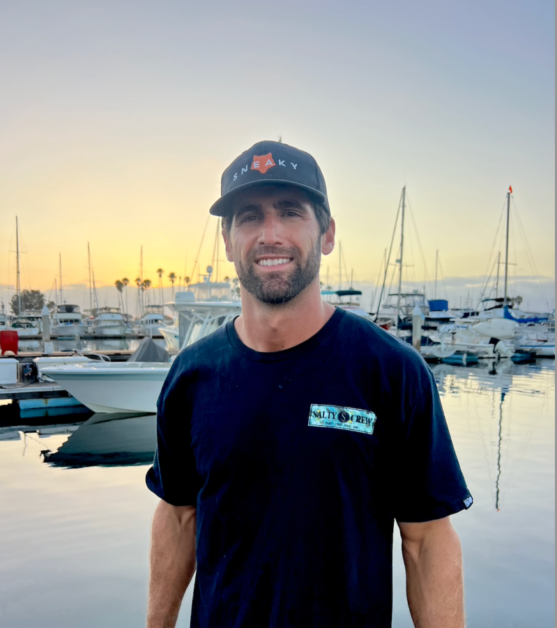 Smiling man with a hat standing in front of boats at sunset.